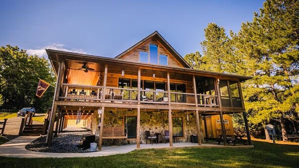 A two-story wooden cabin with a balcony, stone foundation, and a flag. - New River Log Cabin | Waterfront • Sauna • Jet Tub (Pulaski County)