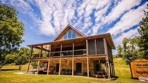 A two-story wooden cabin with a balcony, stone foundation, and a garage. - New River Log Cabin | Waterfront • Sauna • Jet Tub (Pulaski County)