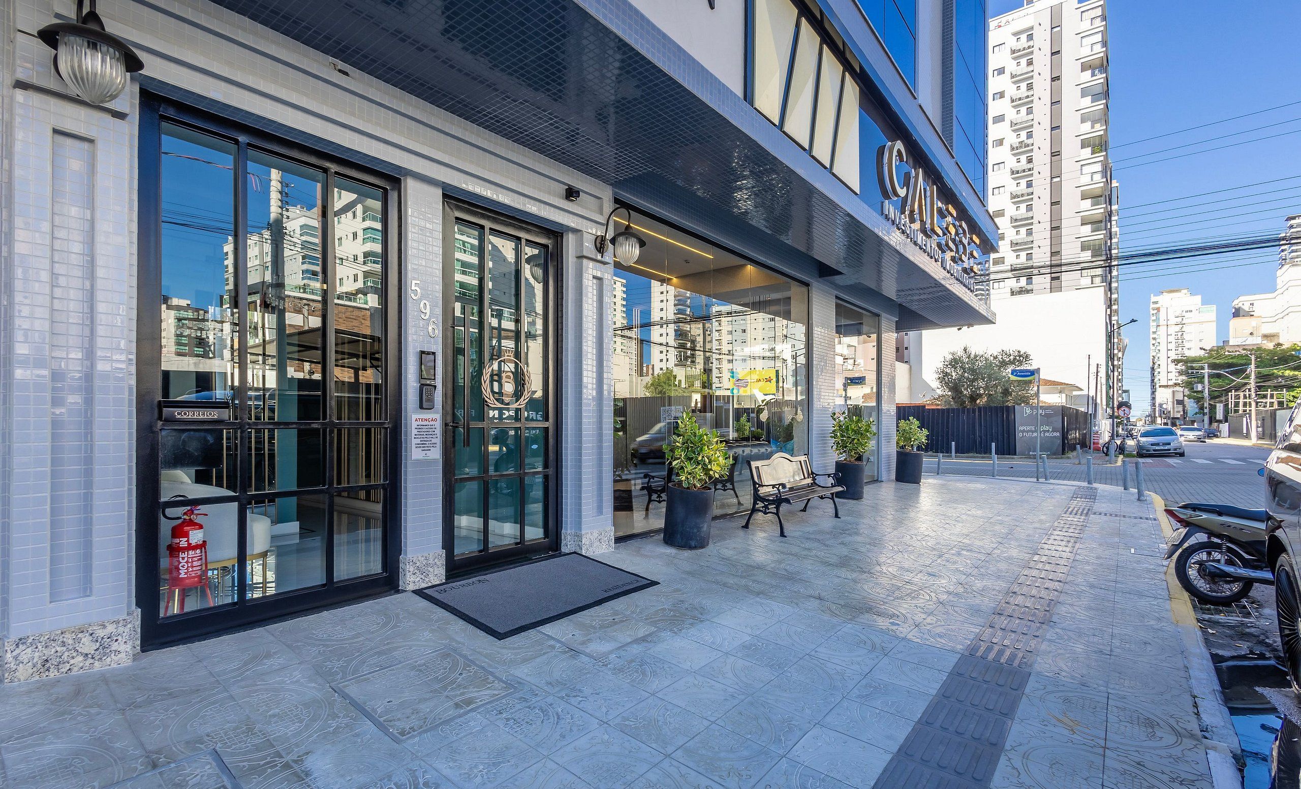 A modern building with large glass windows, a visible entrance, and outdoor seating area with potted plants.