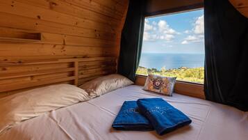 A wooden cabin room with a bed, a window with curtains, and a view of the sea.
