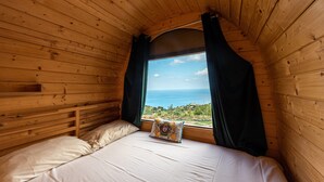 A wooden cabin room with a bed, a window with curtains, and a view of the sea. - Bungalow 'Limone' with Sea View, Wi-Fi and Air Conditioning (Tusa)