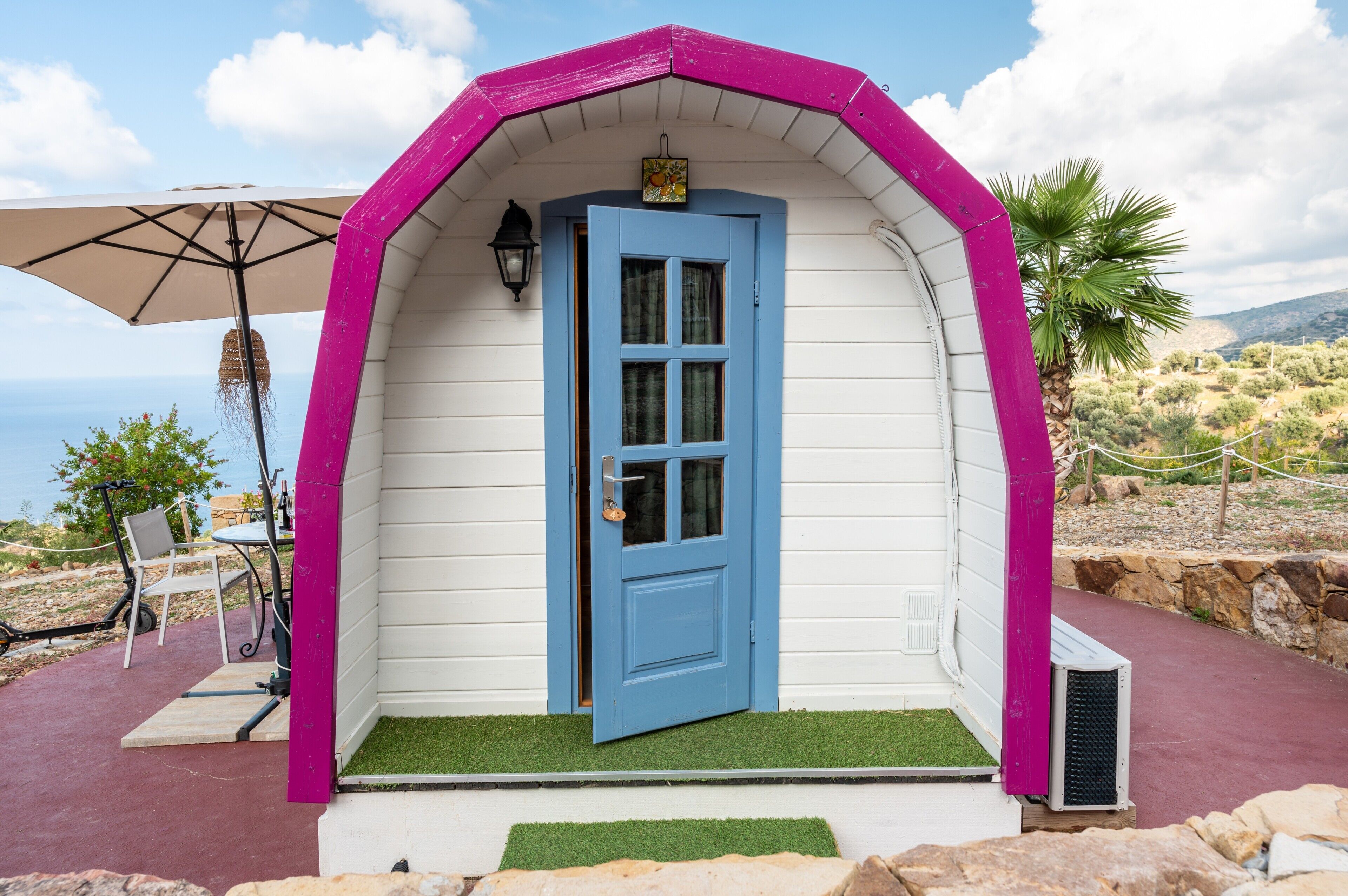 A small, single-story building with a blue door and a pink roof, surrounded by a patio with a table and chairs.
