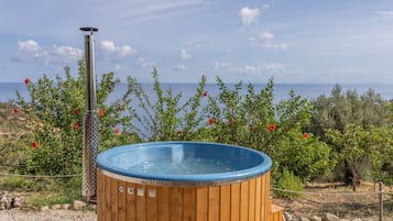 A hot tub with a wooden exterior and a metal heating element, surrounded by greenery and overlooking a body of water.