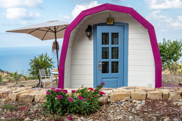 A small, single-story building with a blue door and pink trim, surrounded by a stone wall and a garden with pink flowers.