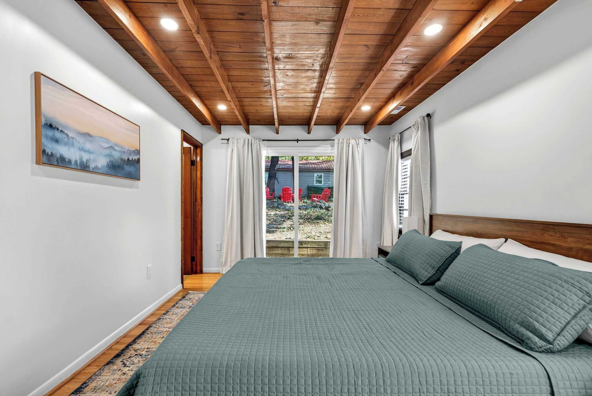 Une chambre avec un grand lit, des poutres apparentes en bois au plafond, un tableau au mur et une vue sur l’extérieur.