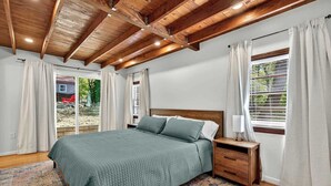 A bedroom with a wooden ceiling, a bed with green bedding, a nightstand, a lamp, and a window with curtains. - Spacious cabin w/private river, hot tub & fire pit (Harpers Ferry)
