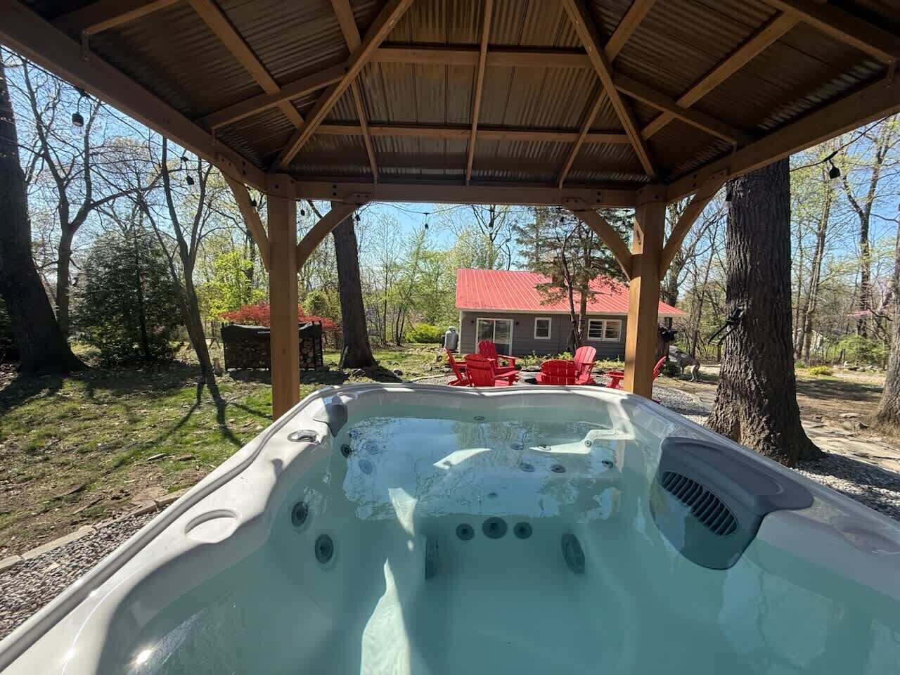 Un bain à remous sous une pergola en bois, avec une maison et des chaises rouges en arrière-plan.