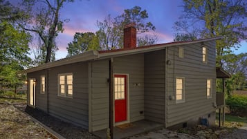 A small, single-story cabin with a red door and multiple windows, surrounded by trees and a stone pathway.