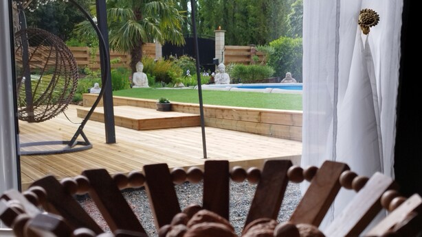 Ein Stuhl aus Holz mit einem besonderen Design, eine Holzterrasse und ein Garten mit Statuen und einem Pool.