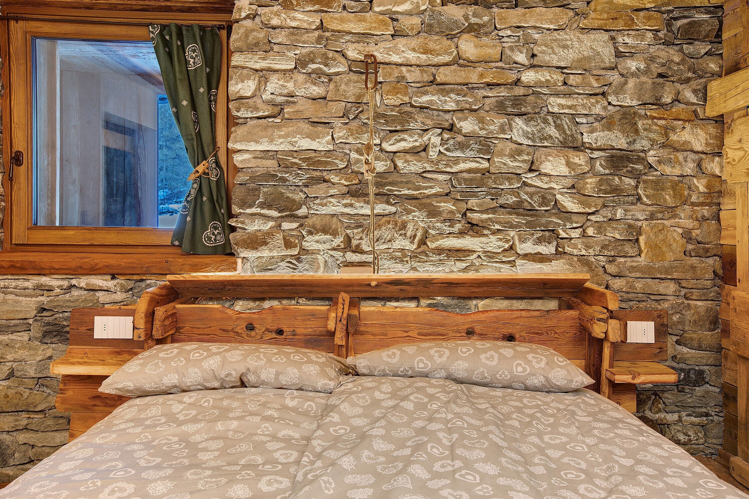 A wooden bed with a headboard against a stone wall, a window with a curtain, and a wooden nightstand with a lamp.