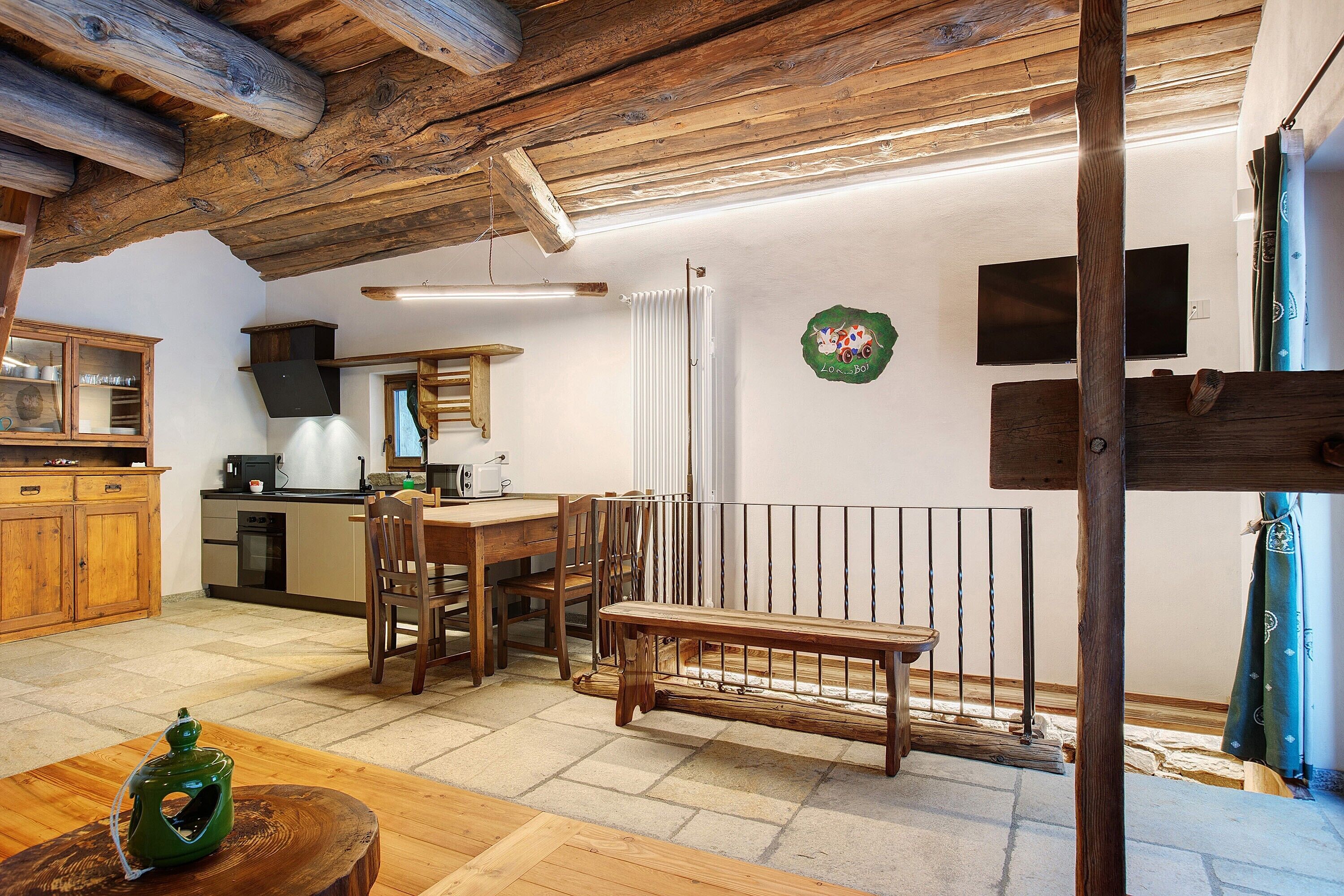 A rustic interior with exposed wooden beams, a dining area with a bench, a kitchen with modern appliances, and a stone floor.