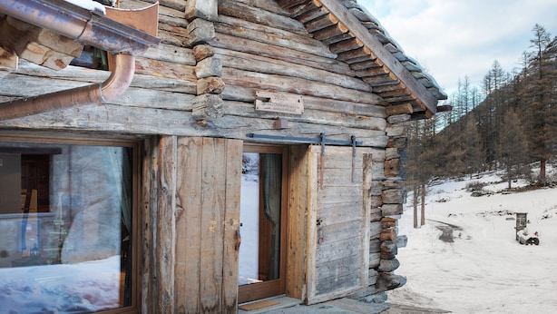A log cabin with a wooden door and large windows, surrounded by snow.