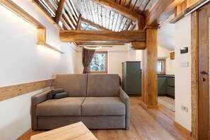 Een gezellige kamer met een bank, houten balken, een raam en een deur naar een keuken.