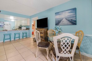 A dining area with a glass table and chairs, a television, and a painting of a pier. - Sugar Beach 132 (Orange Beach)