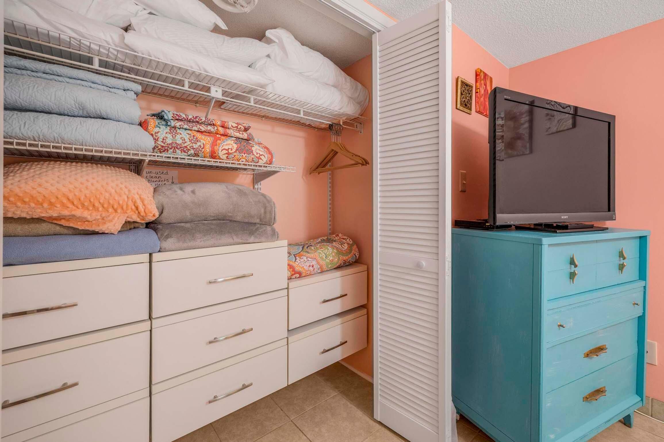 A bedroom with a closet containing folded blankets and pillows, a television on a dresser, and a bed with a white comforter.