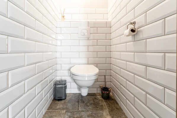 A small, white-tiled bathroom with a toilet, a trash bin, and a bucket.