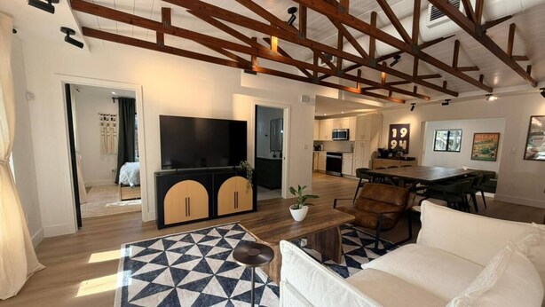A spacious living area with a high ceiling, wooden beams, a large TV, a dining table, and a sofa. - Modern Rustic Miami Retreat | Tree-Shaded Backyard & Fire Pit | Near Wynwood (Miami)
