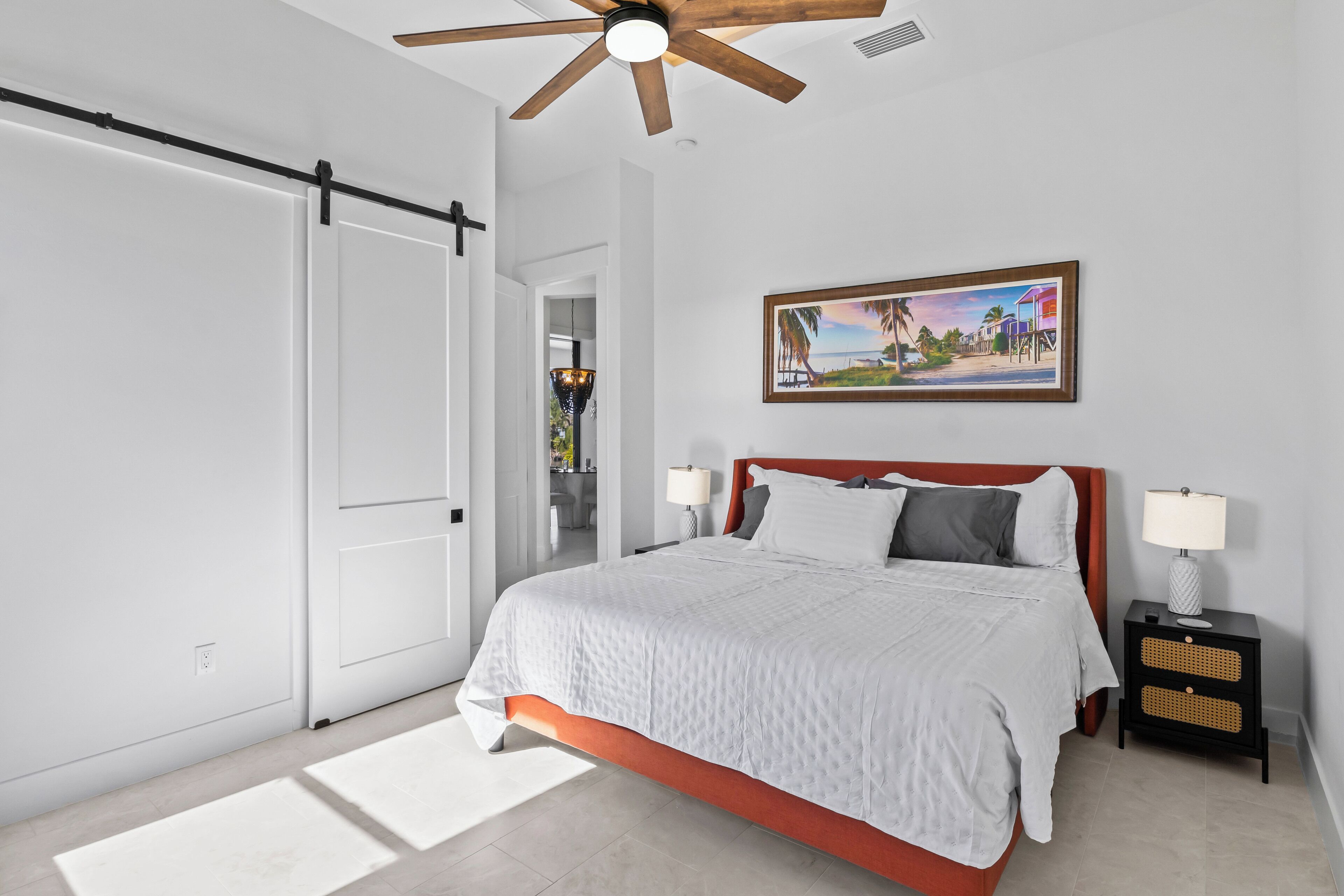 Une chambre à coucher comprenant un lit, une table de chevet, un ventilateur de plafond, une porte et un tableau accroché au mur.