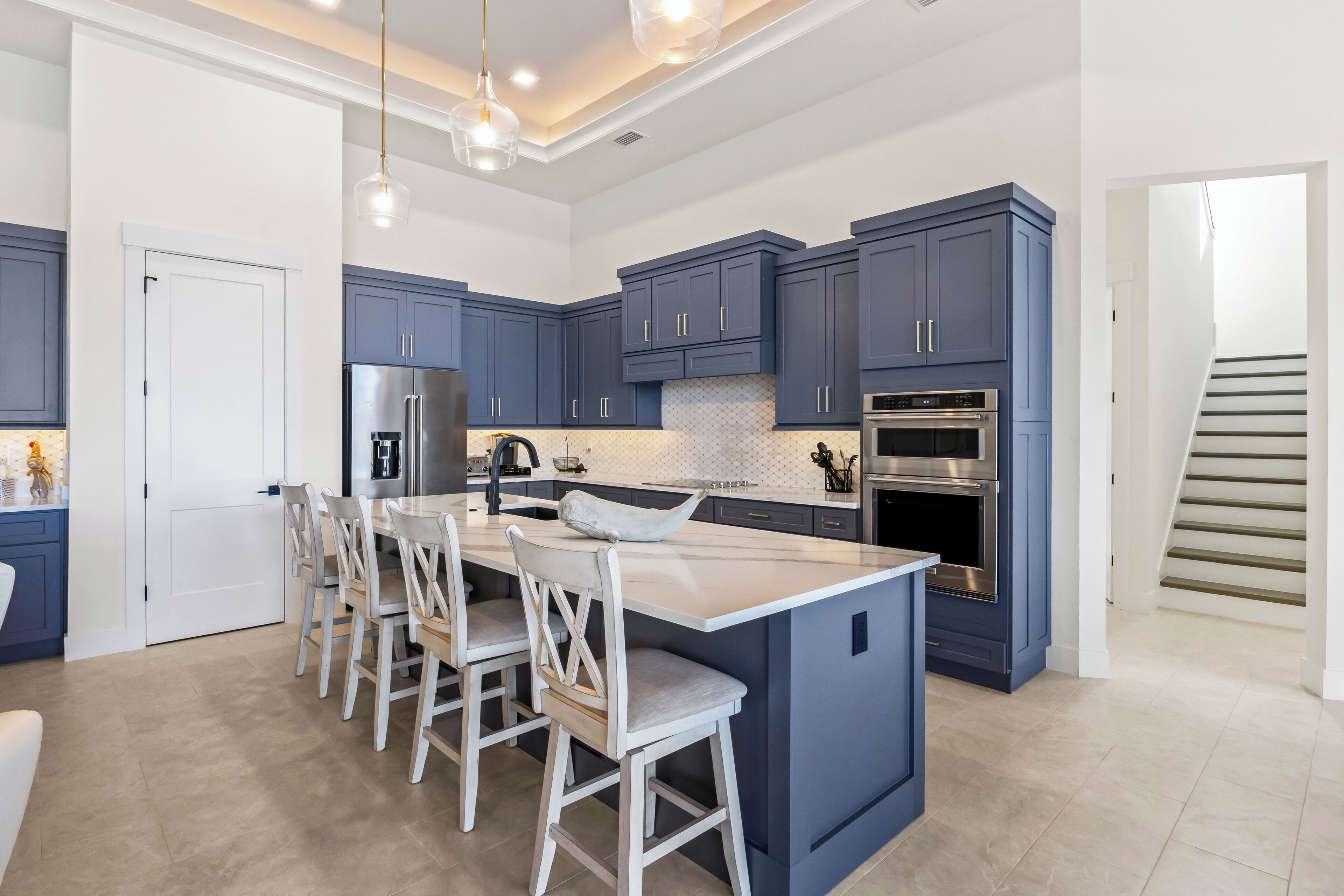 Une cuisine moderne avec des meubles de couleur bleu foncé, un îlot central doté de tabourets de bar, des appareils en acier inoxydable et un éclairage suspendu.