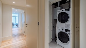 A laundry room with two stacked washing machines, a wooden floor, and a hallway leading to a room with a large window. - Bolhão Prime | Luxe 3BR + Garage (Oporto)