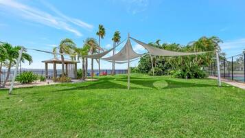 The property features lush tropical gardens with shaded lounging areas, perfect for relaxing outdoors. Palm trees and colorful plants surround the expansive lawn, creating a peaceful, resort-like atmosphere.