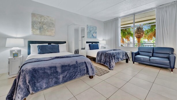 Habitación de hotel con dos camas, un sofá, una mesita de noche y un ventanal con vistas a palmeras.