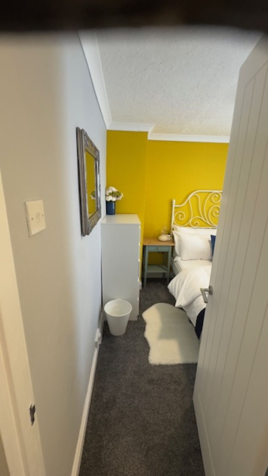 A narrow hallway leading to a bedroom with a yellow accent wall, a white bed, and a small bedside table with a lamp.