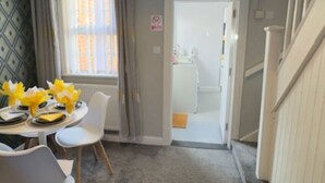 Ruokailutila, jossa on neljälle katettu ruokapöytä, kattokruunu ja näkymä keittiöön oviaukon kautta.
