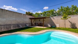 Un patio trasero con piscina, un porche con sillas y una pequeña construcción con techo.