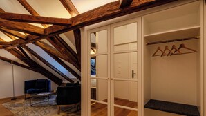 A room with a sloped ceiling, wooden beams, a black chair, a small table, and a white wardrobe with a hanging rod. - Goldener Engl (Hall In Tirol)