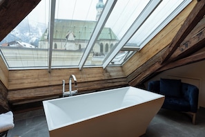Ein modernes Badezimmer mit freistehender Badewanne, Holzbalken und einem Dachfenster mit Blick auf eine Kirche.