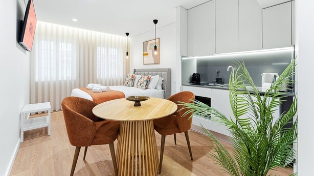Une chambre d’hôtel moderne comprenant un lit, une table de salle à manger ronde avec des chaises, un coin cuisine et une grande plante en pot.