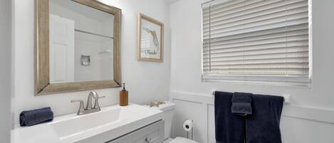Un baño con tocador blanco, un espejo grande, inodoro y ventana con persianas.