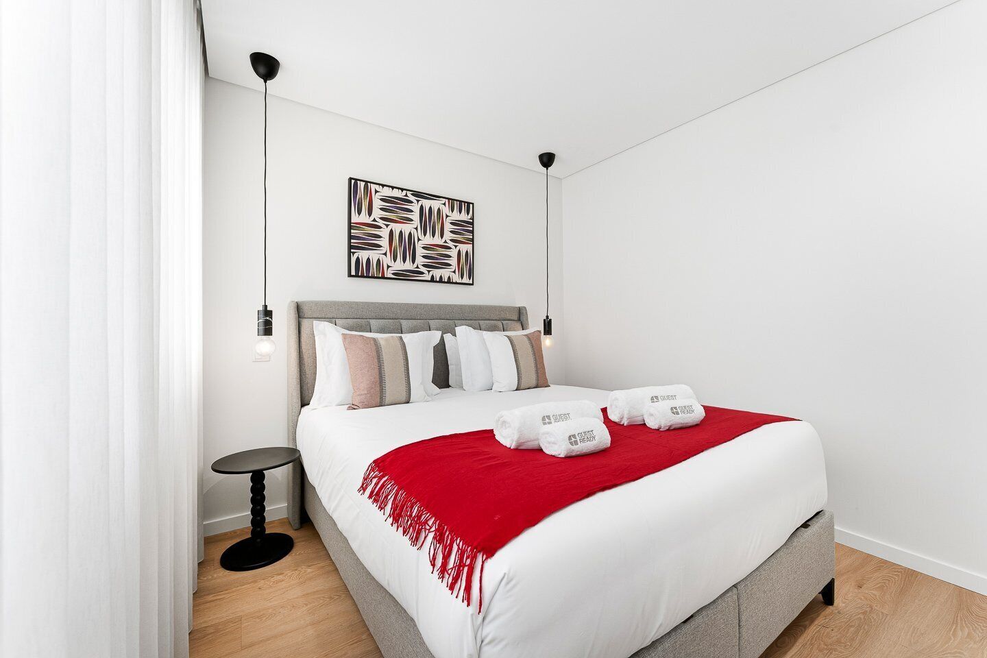 A modern hotel room with a large bed, white bedding, a red throw blanket, and a minimalist decor.