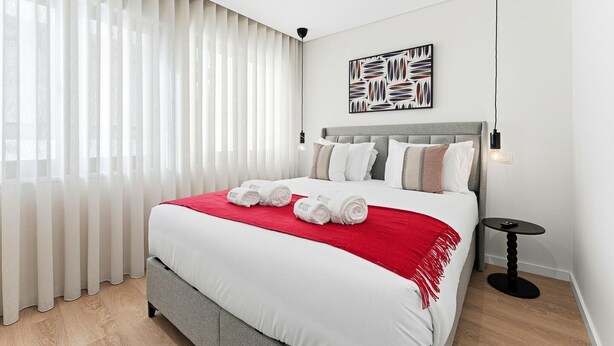 A modern hotel room with a large bed, white bedding, a red throw, and a minimalist decor. - GuestReady The 7 Seas - Caspian Sea 1E (Porto)