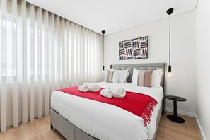 Moderni hotellihuone, jossa on suuri sänky, valkoiset vuodevaatteet, punainen heittopeitto ja pelkistetty sisustus.