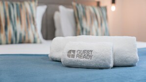 En pænt redt seng med et blåt sengetæppe og et hvidt håndklæde, der er foldet, med broderet 'GUEST READY'.