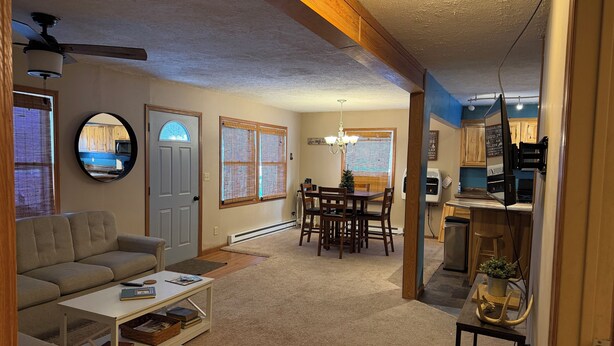 A living room with a sofa, coffee table, and a dining area with a table and chairs. There is a kitchen area with appliances and a refrigerator. - Crossover Cabin is ready for your outdoor adventure! Perfect for snowmobilers!! (Munising)
