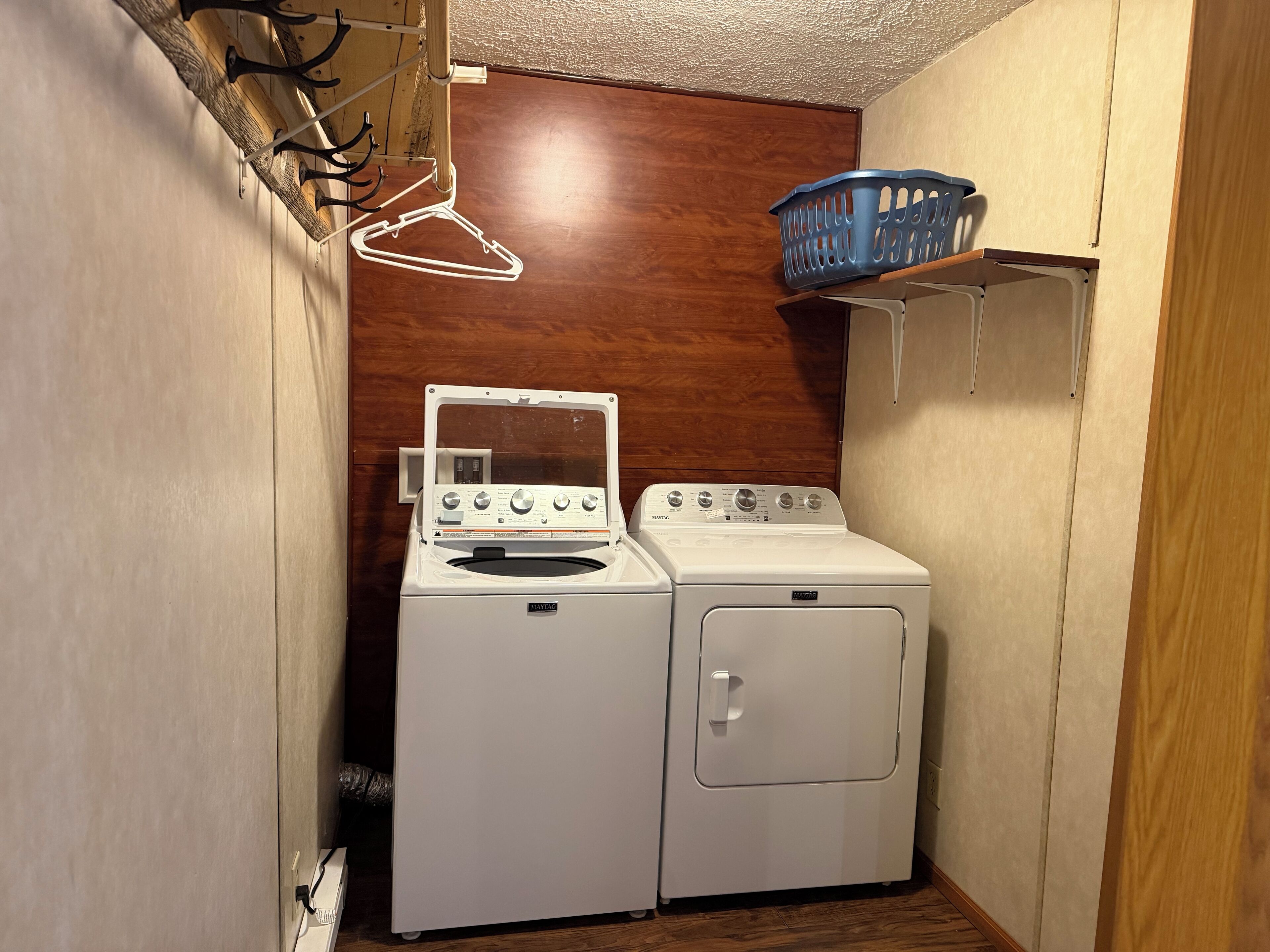 A compact laundry room with a front-loading washer and dryer, wooden flooring, and a blue laundry basket on a shelf.
