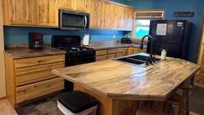 A kitchen with wooden cabinets, a stainless steel microwave, a black stove, a black refrigerator, and a marble countertop island with a sink. - Crossover Cabin is ready for your outdoor adventure! Perfect for snowmobilers!! (Munising)