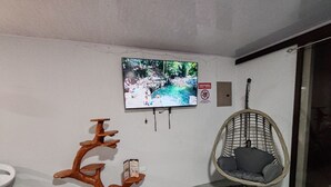 A room with a television mounted on the wall displaying a nature scene, a wicker chair, and a unique wooden stand with multiple shelves. - Modern Aparm with Private Jacuzzi •Arenal Volcano View • 3 Minutes from Downtown (La Fortuna)