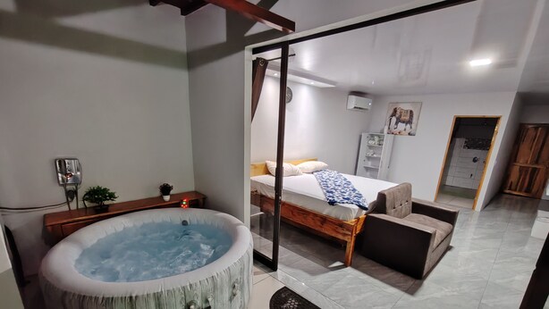 A modern bedroom with a wooden bed, a large hot tub, and a glass door leading to a bathroom. - Modern Aparm with Private Jacuzzi •Arenal Volcano View • 3 Minutes from Downtown (La Fortuna)