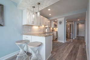 A modern kitchen with white cabinetry, a granite countertop, and pendant lighting. There are two bar stools with woven bases. - 3BR, 2BA Oceanfront Beach Condo w/ 270° Views & Balcony access from every room (Myrtle Beach)