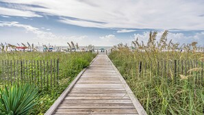 Ein Holzsteg führt zu einem Strand mit Sonnenschirmen und Liegestühlen.