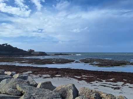 A rocky shoreline with a building on a hill, a clear sky, and calm sea.. “L'Eclose - Les Korrigans” Apartment with Shared Pool, Garden & Wi-Fi – Near GR34