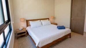 Een keurig opgemaakt bed met witte lakens en een blauwe handdoek, een houten hoofdbord, nachtkastjes met lampen en een groot raam aan de zijkant.