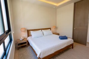 A neatly made bed with white linens and a blue towel, a wooden headboard, bedside tables with lamps, and a large window on one side. - New front beach apartment with golf cart (Puerto Morelos)