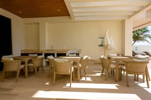 A sunlit outdoor dining area with wooden tables and chairs, a central island with a sink, and a white umbrella. - New front beach apartment with golf cart (Puerto Morelos)