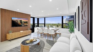 Un salon moderne doté d’un téléviseur à écran plat, d’un coin repas avec une table et des chaises, et de grandes fenêtres offrant une vue sur la ville.