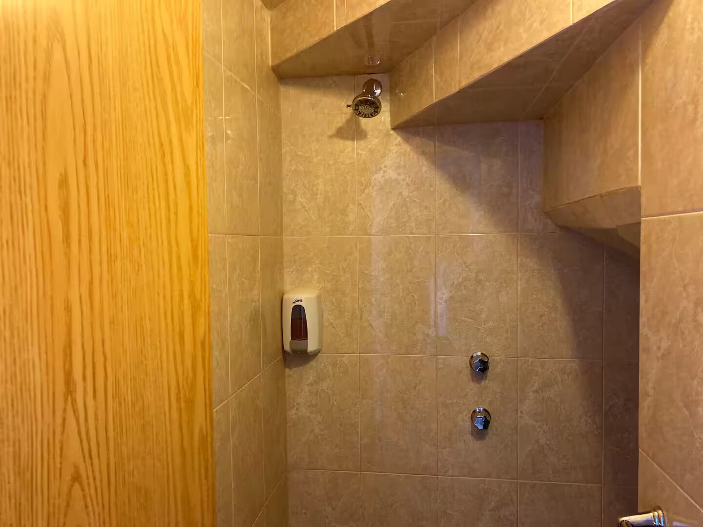 A shower with beige tiles, a soap dispenser, and two faucets.
